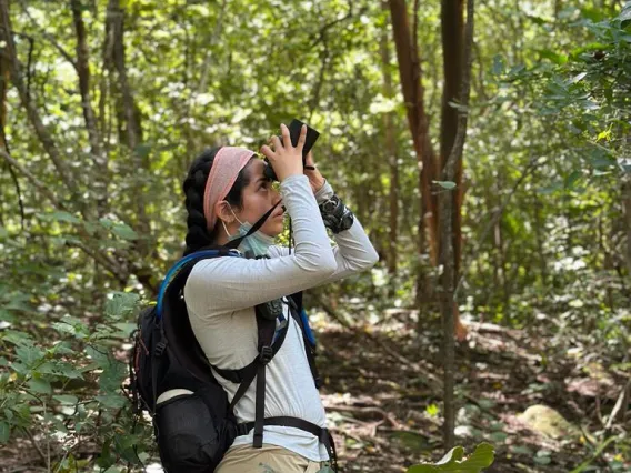 a woman standing in a forest with black hair, a pink headband, a white shirt, and a backpack looking through binoculars