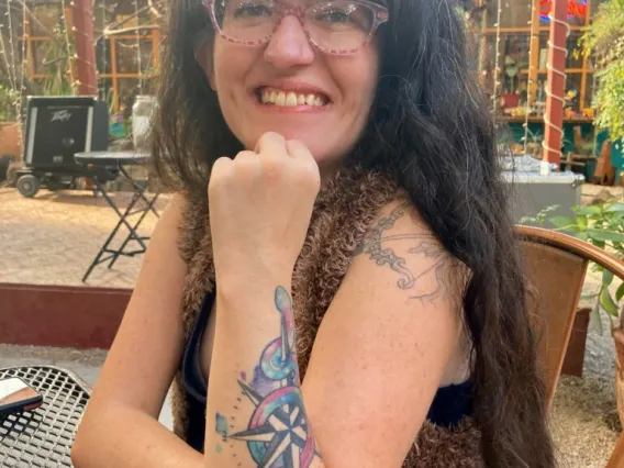 A woman with long black hair and glasses leaning onto a table and smiling