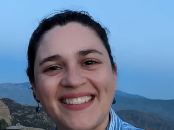 A woman with dark hair pulled back smiling with the sky and mountains behind her