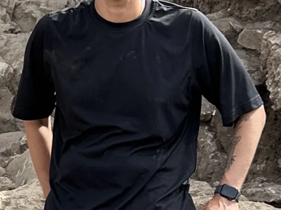 a man with short brown hair wearing a tee shirt and shorts and leaning against a wall of rocks