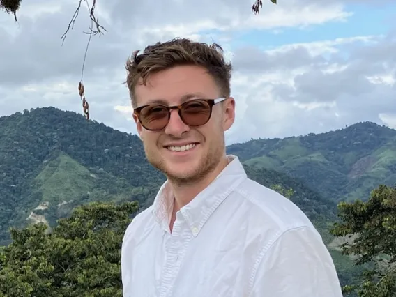 a man with short brown hair and a light colored beard wearing red sunglasses with a forest and mountain in the background