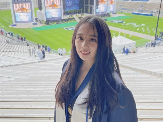 A woman with chest length brown hair smiling while wearing graduation robes, stoles, and a meal standing in the bleachers of a football stadium