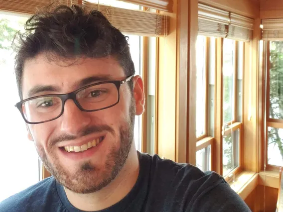 A photo of a man with a beard, glasses and short brown hair smiling with windows in the background