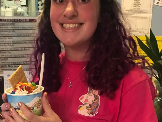 A white woman with wavy brown hair wearing a red shirt with a unicorn on it is holding a cup of ice cream with the background being within the ice cream store.