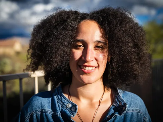 a young black woman smiles at the camera, with shoulder length black hair. In the background a blury sky and desert can be seen.