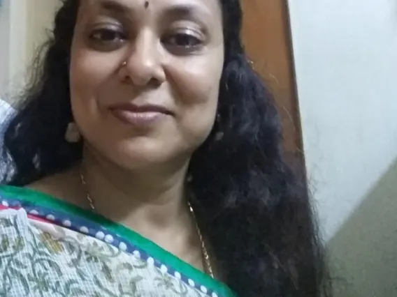 An Indian woman with long black hair wearing a sari, a golden necklace, and a bindi on her forehead. She is standing in front of a wall.