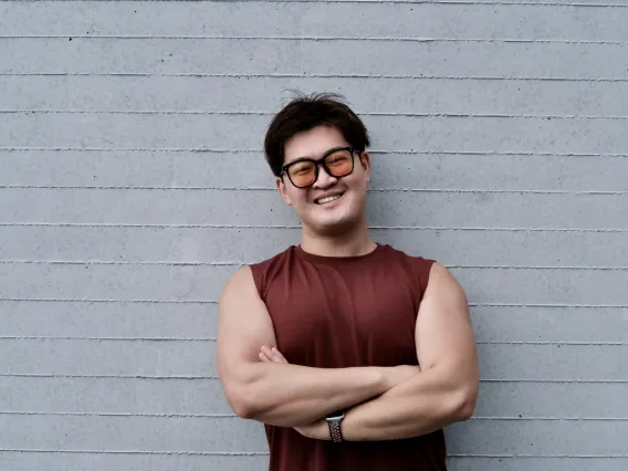 A young east asian man with short black hair is smiling while standing against a grey brick wall. He is wearing a sleeveless red shirt, a watch, and orange tinted sunglasses.