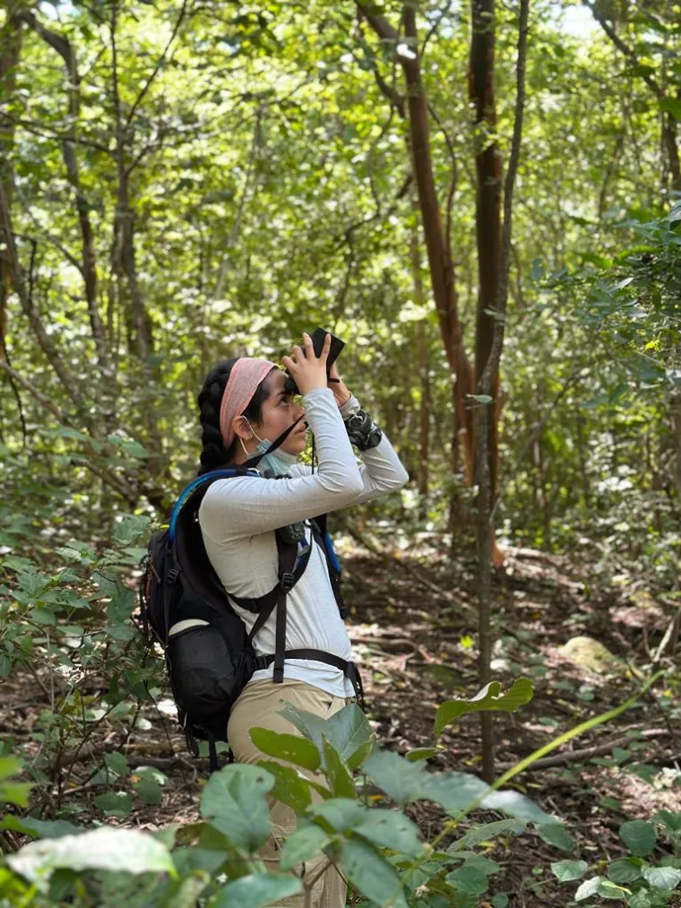 a woman standing in a forest with black hair, a pink headband, a white shirt, and a backpack looking through binoculars