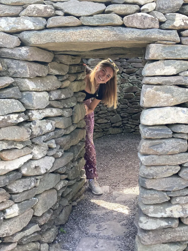 A woman with long blend hair peaking through a doorway of a structure made from stacked stone bricks