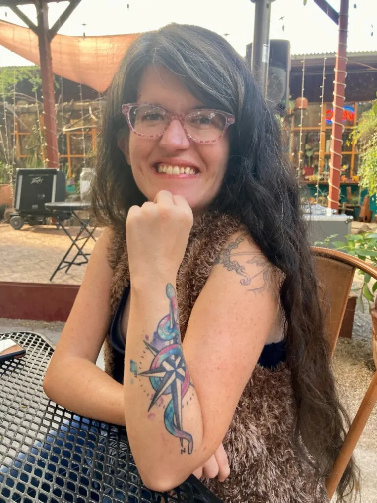 A woman with long black hair and glasses leaning onto a table and smiling