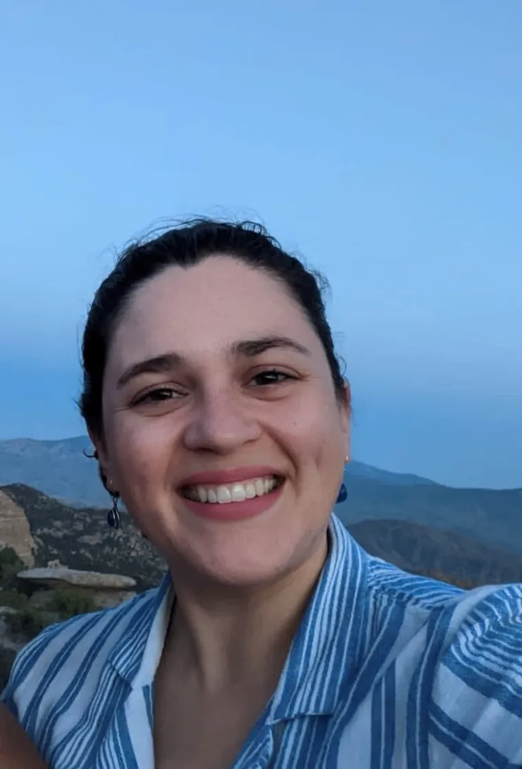 A woman with dark hair pulled back smiling with the sky and mountains behind her