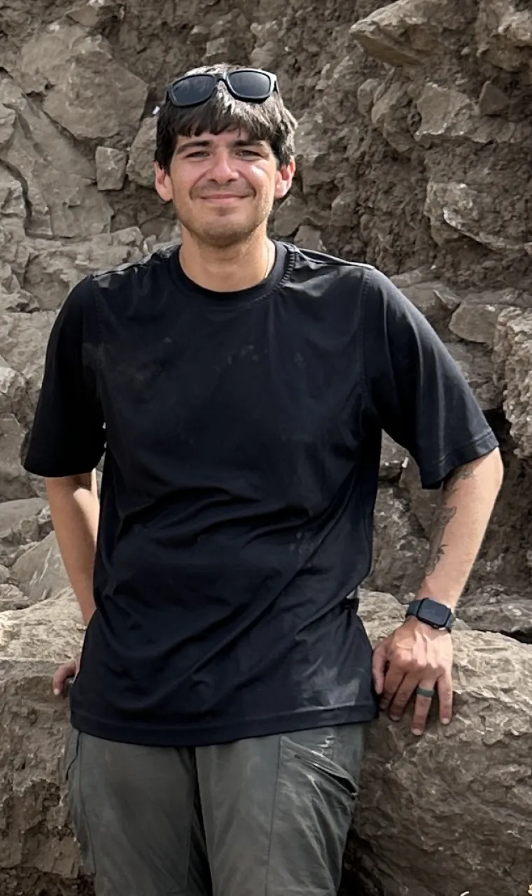 a man with short brown hair wearing a tee shirt and shorts and leaning against a wall of rocks