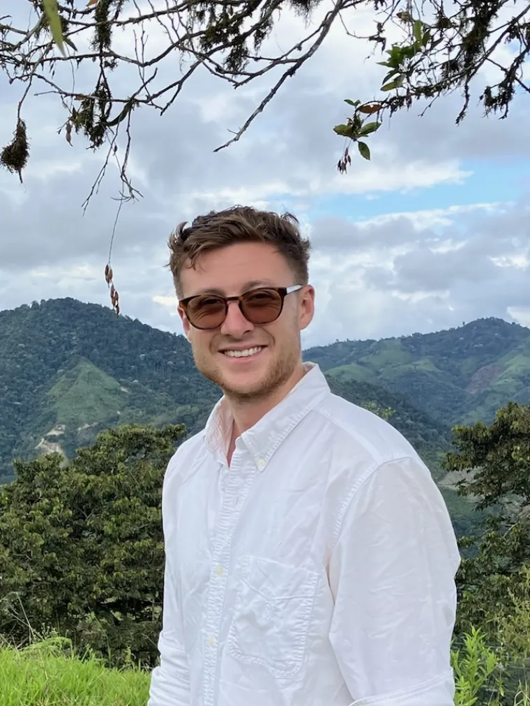 a man with short brown hair and a light colored beard wearing red sunglasses with a forest and mountain in the background