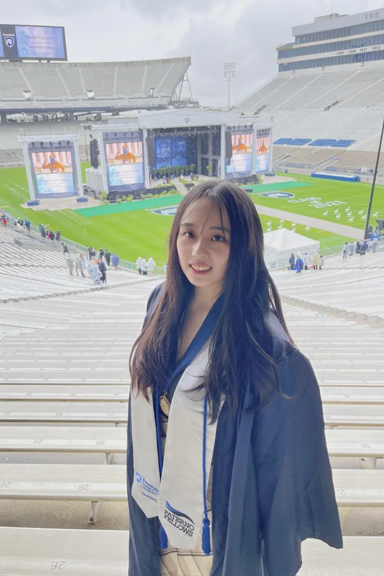 A woman with chest length brown hair smiling while wearing graduation robes, stoles, and a meal standing in the bleachers of a football stadium