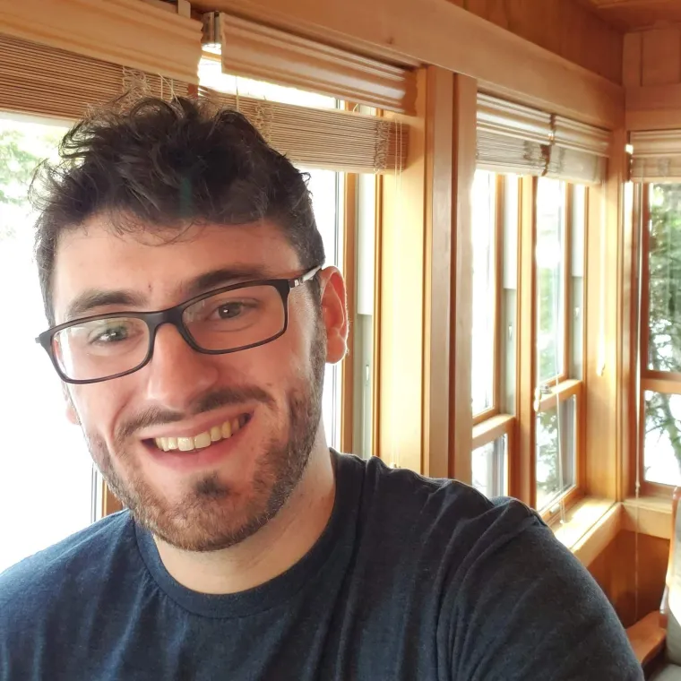 A photo of a man with a beard, glasses and short brown hair smiling with windows in the background