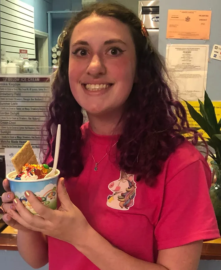 A white woman with wavy brown hair wearing a red shirt with a unicorn on it is holding a cup of ice cream with the background being within the ice cream store.