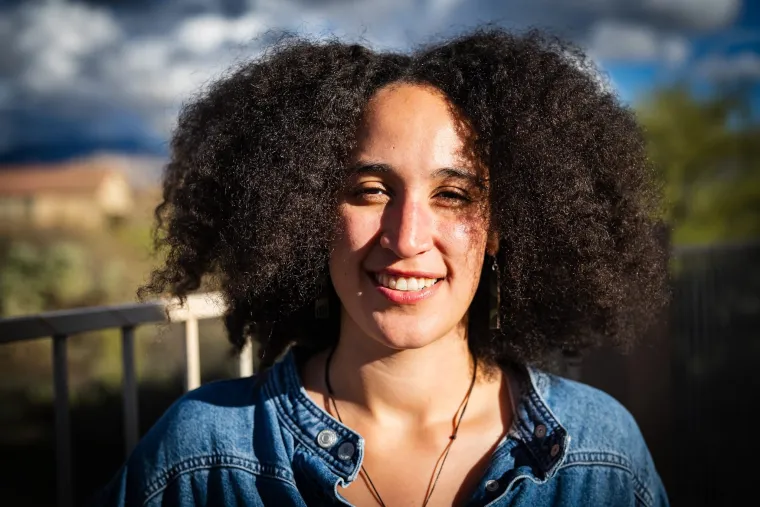 a young black woman smiles at the camera, with shoulder length black hair. In the background a blury sky and desert can be seen.