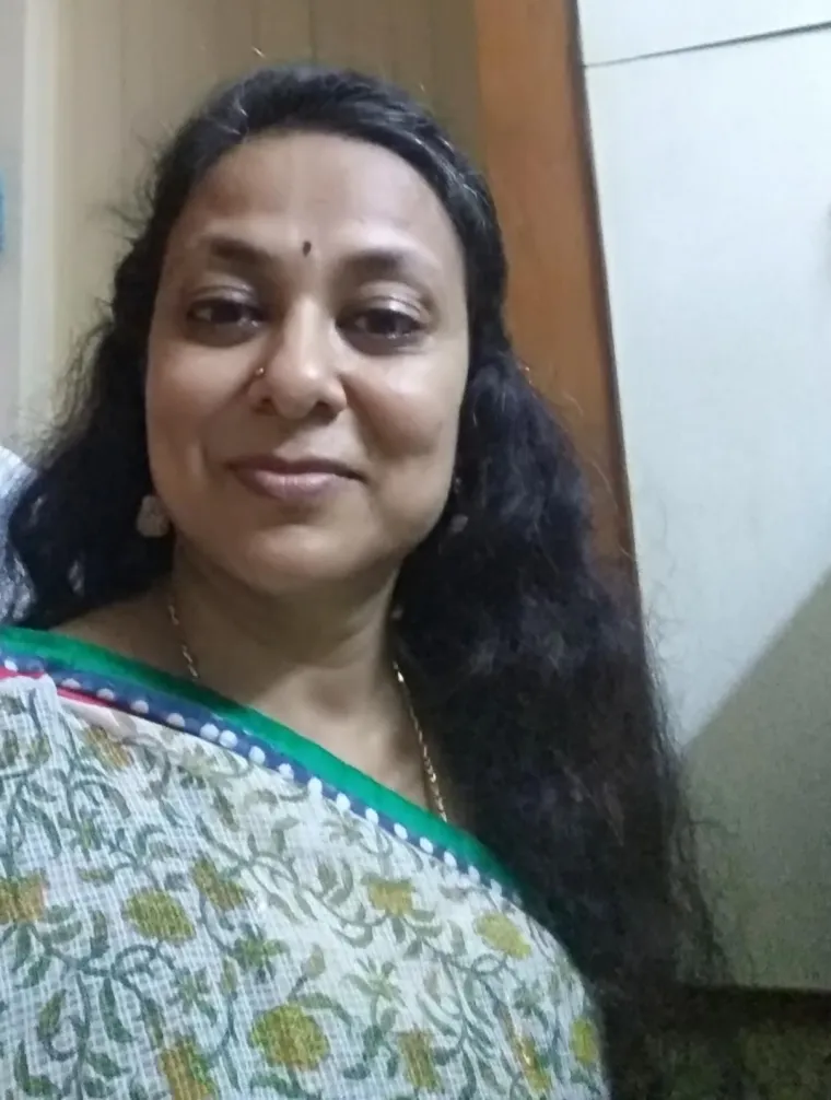 An Indian woman with long black hair wearing a sari, a golden necklace, and a bindi on her forehead. She is standing in front of a wall.