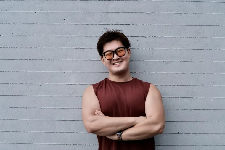 A young east asian man with short black hair is smiling while standing against a grey brick wall. He is wearing a sleeveless red shirt, a watch, and orange tinted sunglasses.