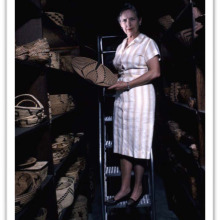 woman standing on ladder in storage room full of baskets, holding a basket
