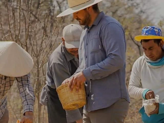 In the center of the picture is a tall hispanic man in a wide brimmed hat with a dark beard he is holding a basket in his hands. Three older hispanic men is similair clothes hold smaller baskets. They all stand in a desert environment.