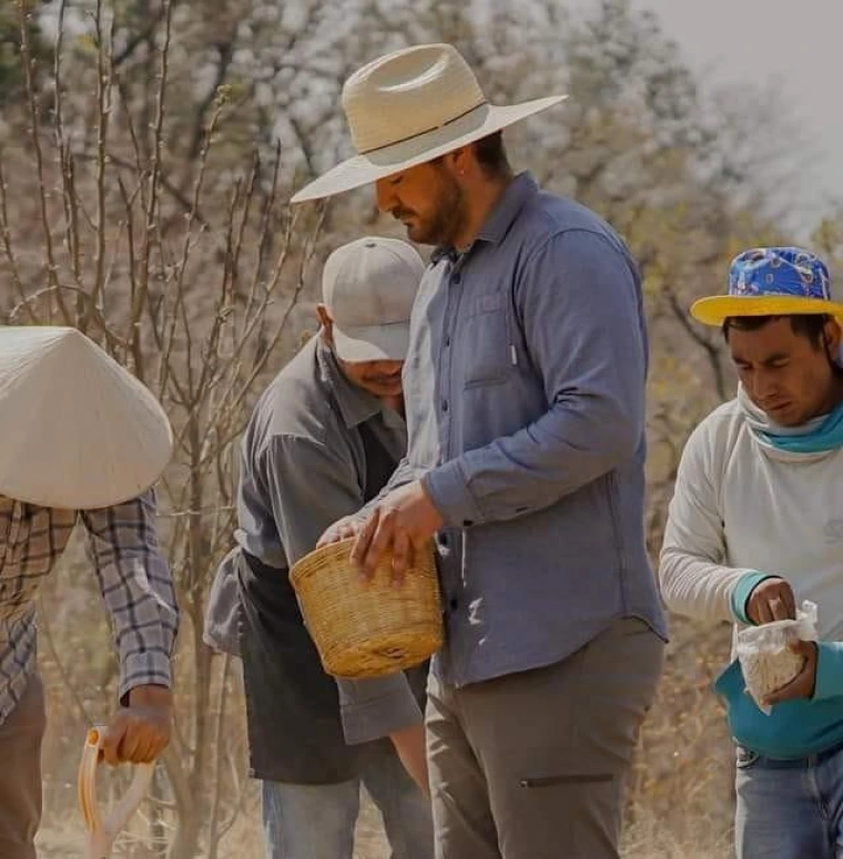 In the center of the picture is a tall hispanic man in a wide brimmed hat with a dark beard he is holding a basket in his hands. Three older hispanic men is similair clothes hold smaller baskets. They all stand in a desert environment.