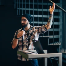Bearded man in black knit cap holding microphone, one arm upraised